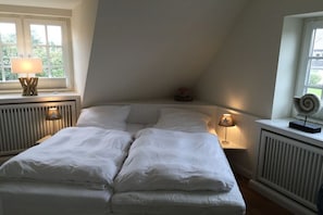A bedroom with a bed, two bedside lamps, a radiator, and a window with a view of greenery. - Holiday home with 1 bedroom (Westerland)