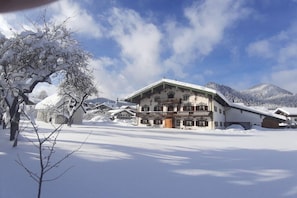 A snow-covered building with a balcony, surrounded by snow-covered trees and mountains. - Landhaus Pichler holiday apartment Kienberg (Ruhpolding)