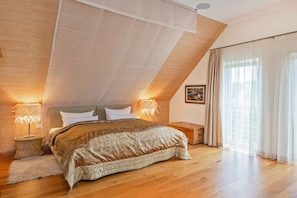 A bedroom with a sloped ceiling, a bed with a beige blanket, two bedside lamps, a wooden nightstand, a small rug, and a window with sheer curtains. - Holiday apartment with 1 bedroom (Rankwitz)