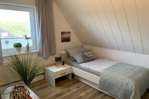 A bedroom with a sloped ceiling, a bed with a grey blanket, a bedside table with a lamp, a window with a view of a green landscape, and a small plant on the windowsill. - Wadden Sea walkers - calm winds (Wurster Nordseeküste)