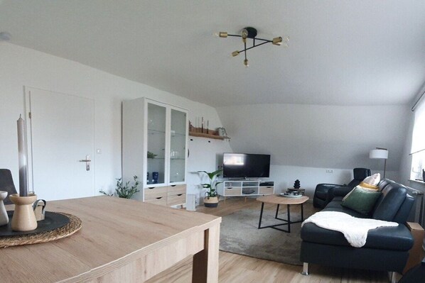 A modern living room with a wooden dining table, a black sofa, a television, and a ceiling light fixture. - Holiday apartment with 2 bedrooms (Weißenstadt!)