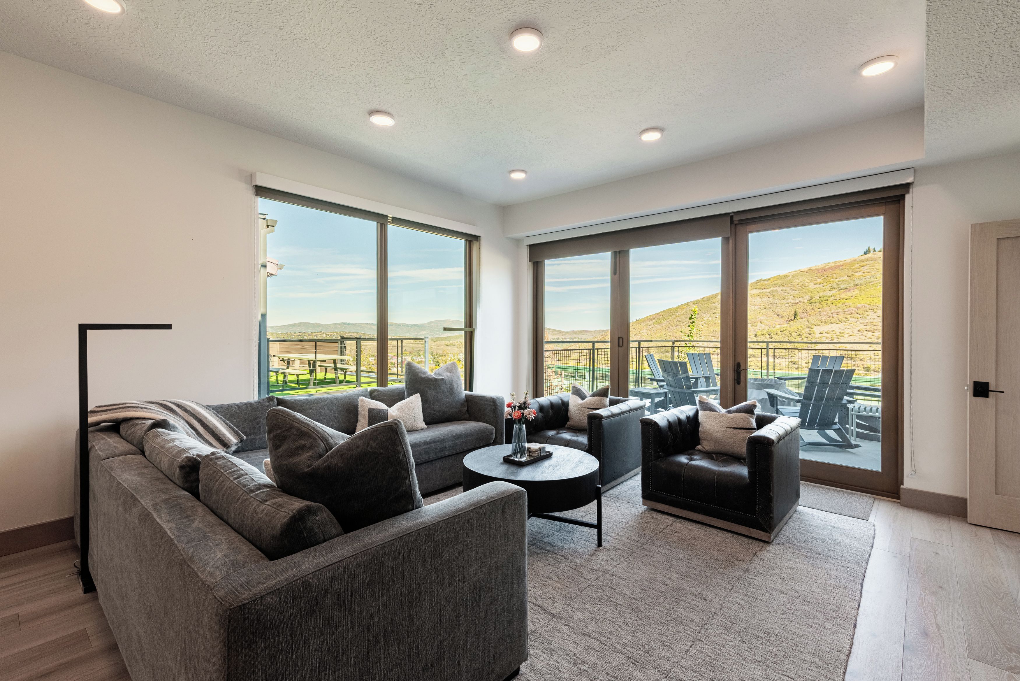 A modern living room with a grey sofa, armchairs, and a coffee table. Large windows offer a view of a hilly landscape.