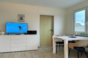A modern room with a flat-screen TV on a white cabinet, a wooden door, a dining table with two chairs, and a window with blinds. - House by the Lighthouse - Holiday Apartments (Neuharlingersiel)