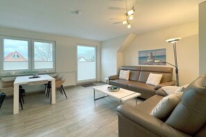 A modern living room with a sofa, coffee table, and dining area. There are large windows and a ceiling fan. - House by the Lighthouse - Holiday Apartments (Neuharlingersiel)