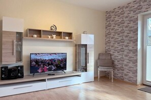 A modern living room with a flat-screen TV, built-in shelves, a wicker chair, and a brick-patterned wall. - House by the Lighthouse - Holiday Apartments (Neuharlingersiel)