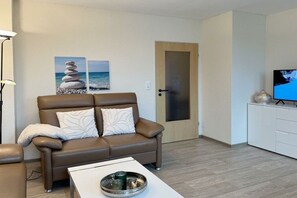 A modern living room with a brown leather sofa, a white coffee table, and a television on a white cabinet. - House by the Lighthouse - Holiday Apartments (Neuharlingersiel)