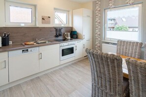 A compact kitchen with a dishwasher, stove, and a coffee maker. There are wicker chairs and a window providing a view of the outside. - House by the Lighthouse - Holiday Apartments (Neuharlingersiel)