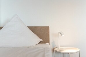 A bed with a white pillow and a headboard, a bedside table with a lamp, and a plain wall. - Holiday apartment with 1 bedroom (Norderney)