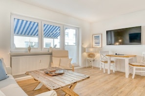 A room with a wooden coffee table, a sofa, a dining table with chairs, and a large window with a striped awning. - Holiday apartment with 1 bedroom (Norderney)