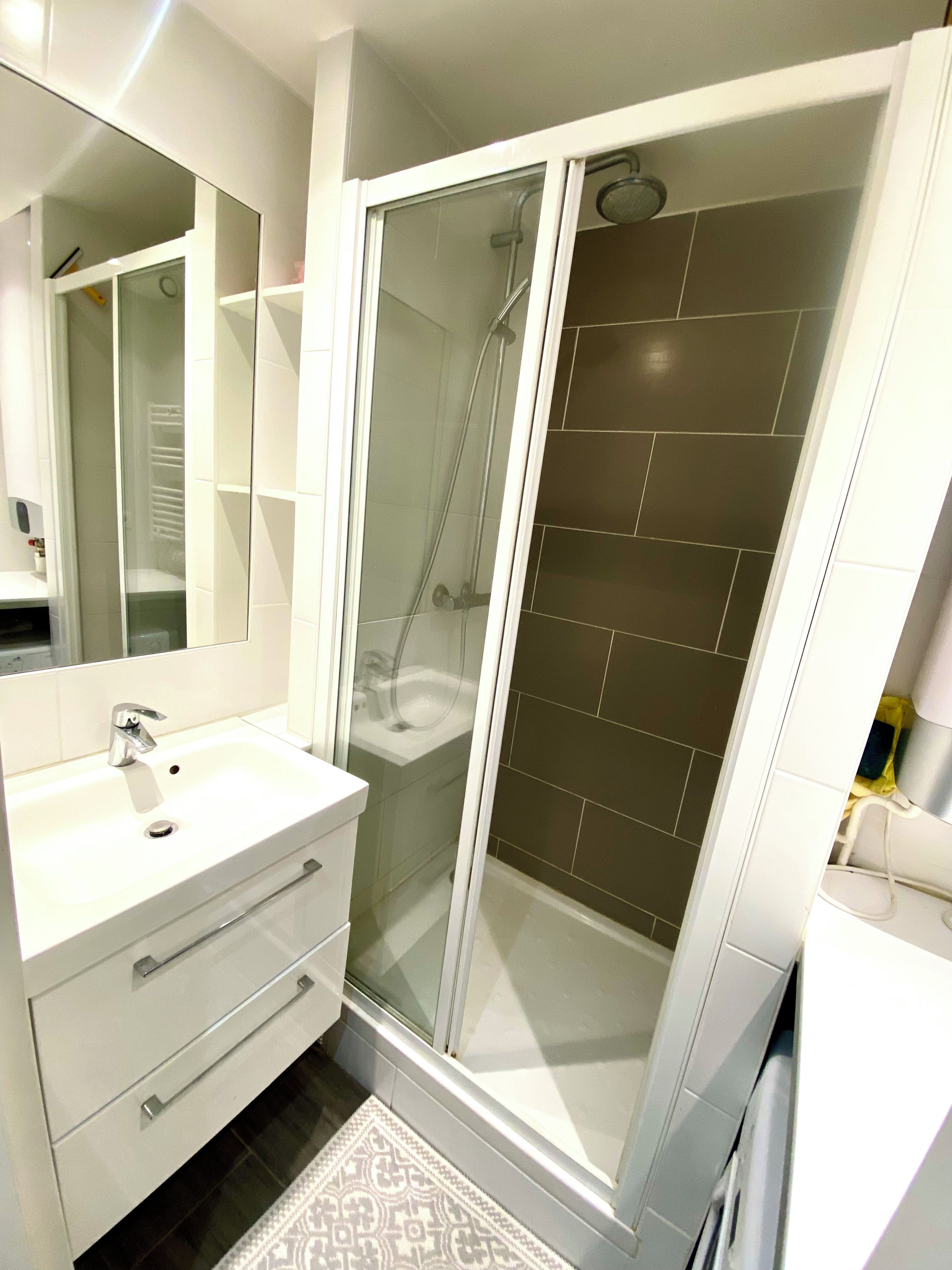 Une salle de bain moderne avec une douche à parois de verre, un lavabo blanc avec un miroir et un tapis à motifs.