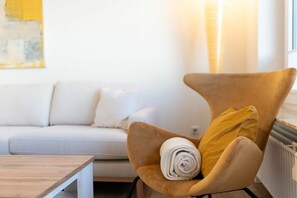 A modern living room with a sofa, a wooden coffee table, and a unique curved armchair with a rolled-up towel and a yellow pillow. - Holiday apartment with 1 bedroom (Norderney)