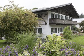Un bâtiment à deux étages doté d’un balcon, entouré d’un jardin où poussent différentes plantes et fleurs.