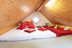 Ein kleines, gemütliches Schlafzimmer im Dachgeschoss mit schräger Decke, einem Fenster und einem Bett mit rot-weißer Bettwäsche. Auf einem roten Teppich liegen mehrere Stofftiere.