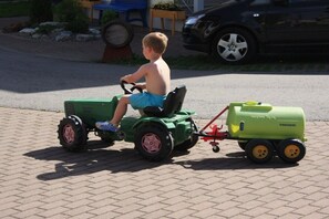 Ein Kind fährt mit einem grünen Spielzeugtraktor mit Anhänger auf einer asphaltierten Fläche.