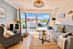 A modern living room with a sofa, armchair, and coffee table, a television, and a view through a glass door to the outside. - ||----------------------Beachcomber F4---------------------|| (Norderney)