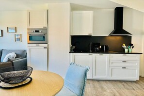 A compact kitchen with white cabinetry, a black range hood, and a dining area with a round table and a chair. - ||----------------------Beachcomber F4---------------------|| (Norderney)
