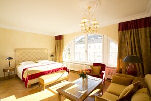 A hotel room with a bed, a sitting area with a sofa and armchair, a coffee table, and a large window with curtains. - Moknis Palais Hotel and Spa (Bad Wildbad)