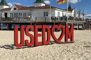 Een strand met een groot, rood bord waarop 'USEDOM' staat, en een gebouw met een groen dak op de achtergrond.