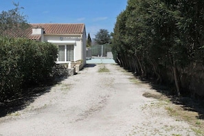 Un chemin de gravier mène à un bâtiment blanc surmonté d’un toit en tuiles, le tout étant entouré d’un feuillage dense.