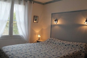 Une chambre à coucher avec un lit, une table de chevet avec une lampe, une fenêtre avec des rideaux et un luminaire fixé au mur.
