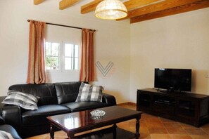 A living room with a black leather sofa, a wooden coffee table, a television on a dark wooden stand, and a window with curtains. - Finca Tugores [1970A] (Lloseta)