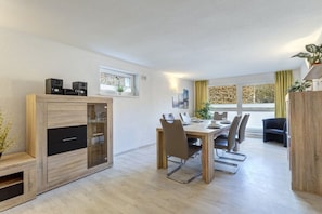 A modern dining area with a wooden table and chairs, a built-in cabinet with speakers, and a view of the outdoors through large windows. - Holiday apartment with 2 bedrooms (Seebad Ahlbeck)