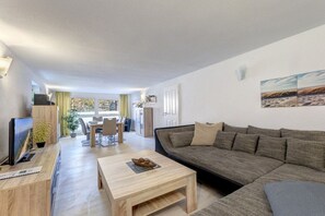 A modern living room with a sofa, coffee table, and a television. There is a dining area with chairs and a table in the background. - Holiday apartment with 2 bedrooms (Seebad Ahlbeck)