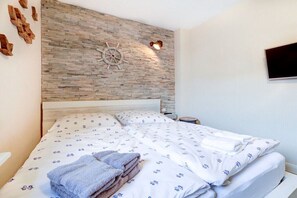A neatly made bed with a patterned bedspread and pillows, a wooden headboard, a wall-mounted ship's wheel decoration, and a flat-screen TV on the wall. - Villa by the sea Apartment 4 (St. Peter-Ording)