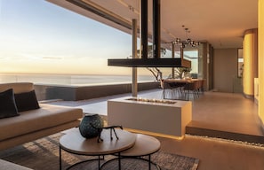 A modern living room with a view of the sea, a fireplace, and a dining area. - Epic Villa by Golden Sands Escape (Cape Town)