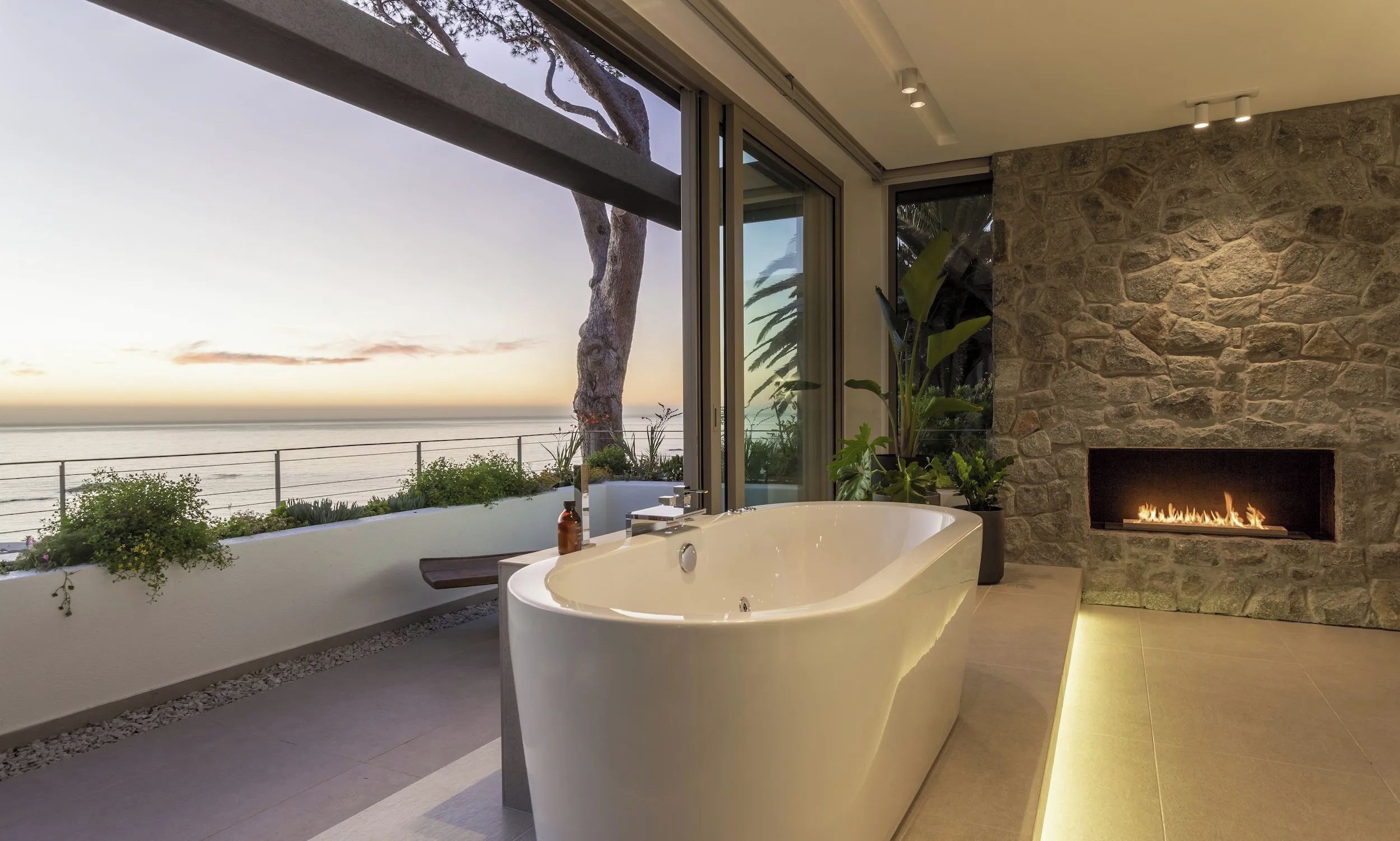 A modern bathroom with a large bathtub, a stone fireplace, and a view of the ocean.
