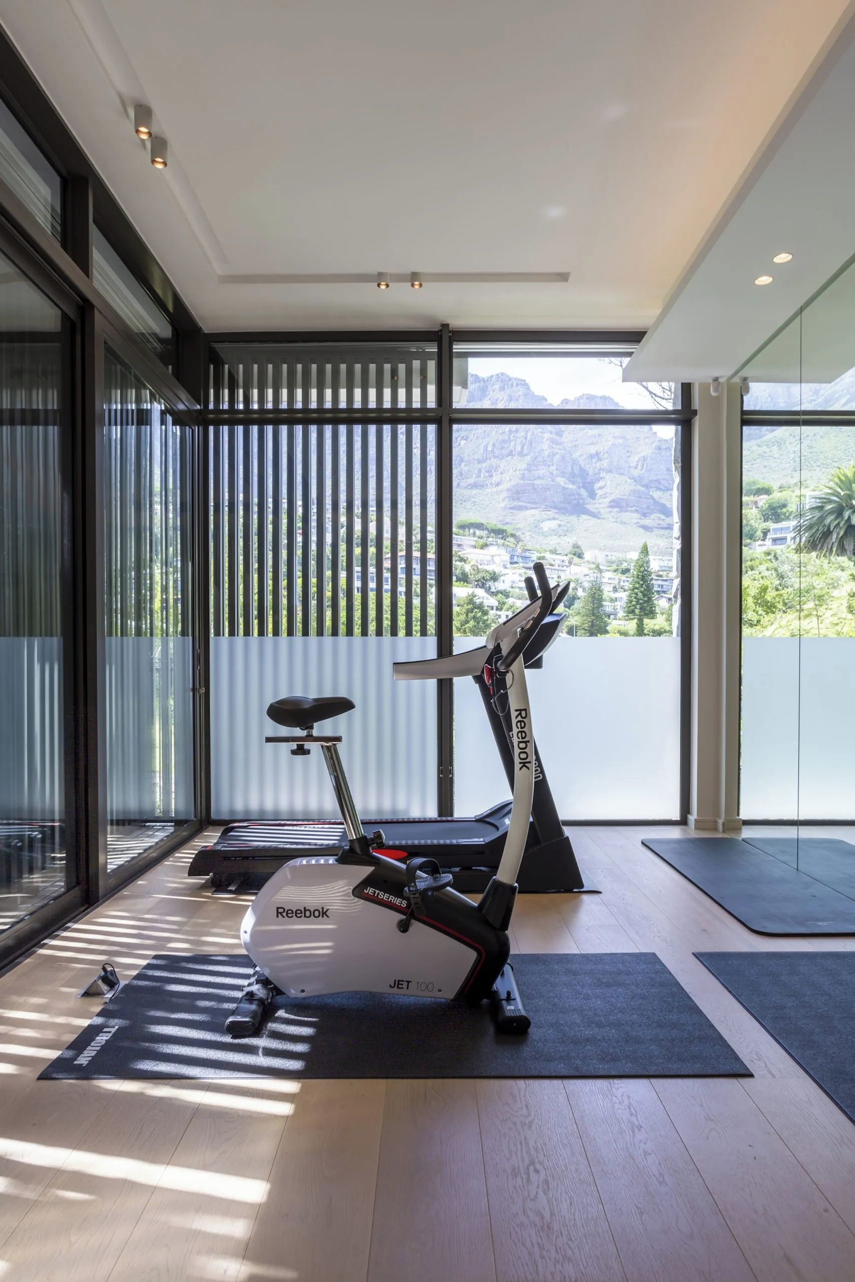 Un gimnasio moderno en casa con bicicleta estática y cinta de correr, amplias ventanas con vista a la montaña, y piso de madera.