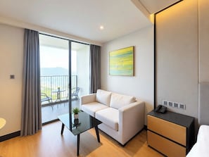 A modern hotel room with a sofa, a glass table with a plant, a bedside table with a phone, and a view of the outdoors through a sliding door. - Soliel Hotel Da Nang (An Hai)