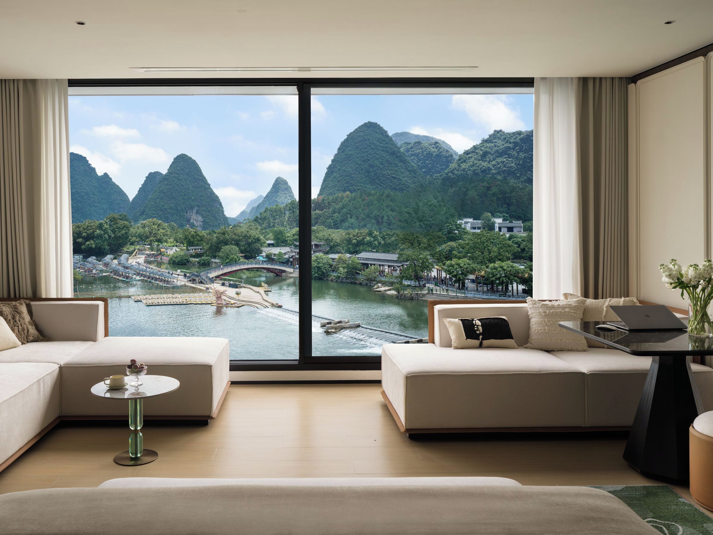 A modern hotel room with a large window offering a view of a river and mountains. There are two sofas and a small table with a laptop.
