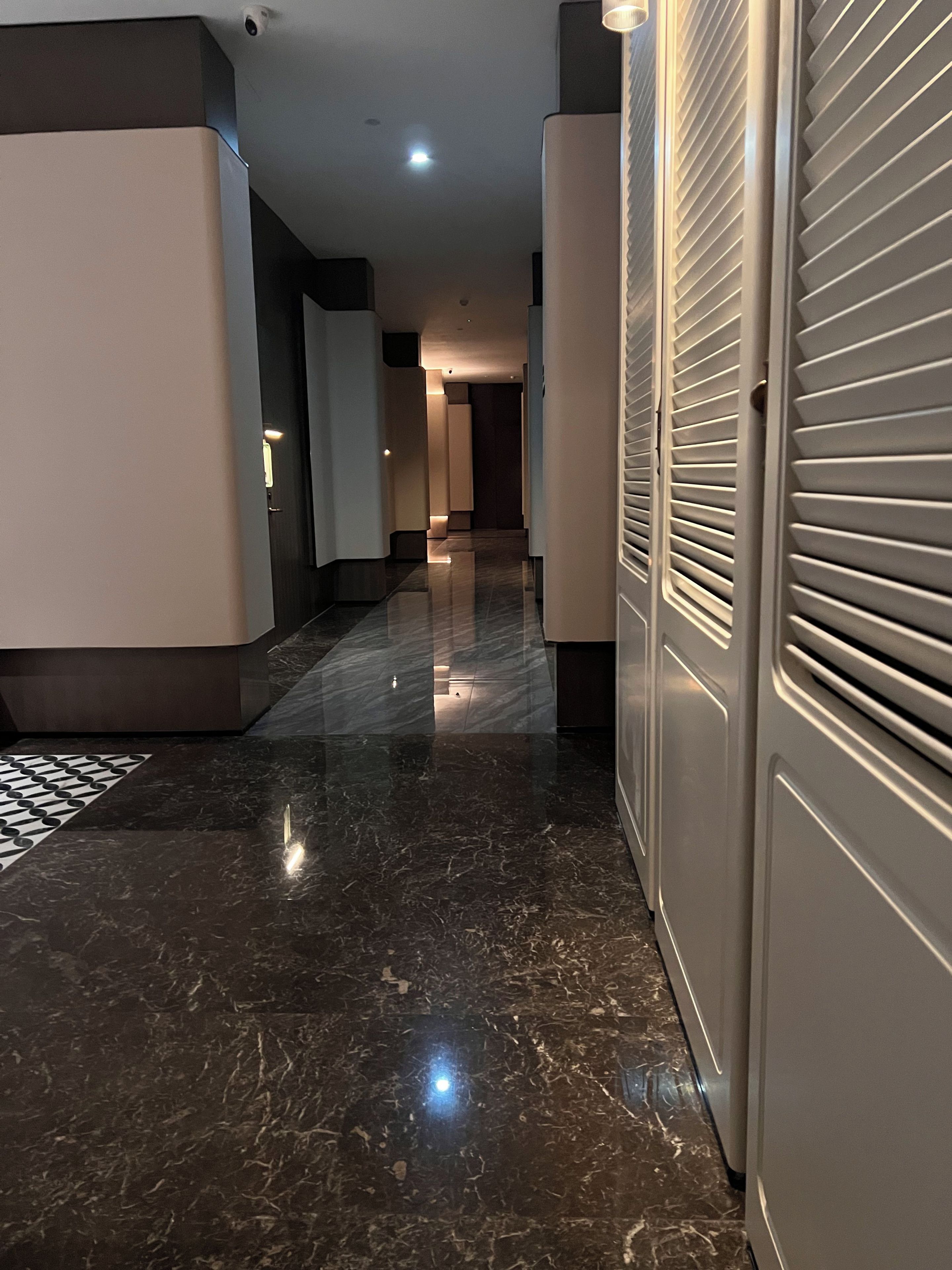 A corridor with a shiny marble floor, white walls, and a door with horizontal slats.