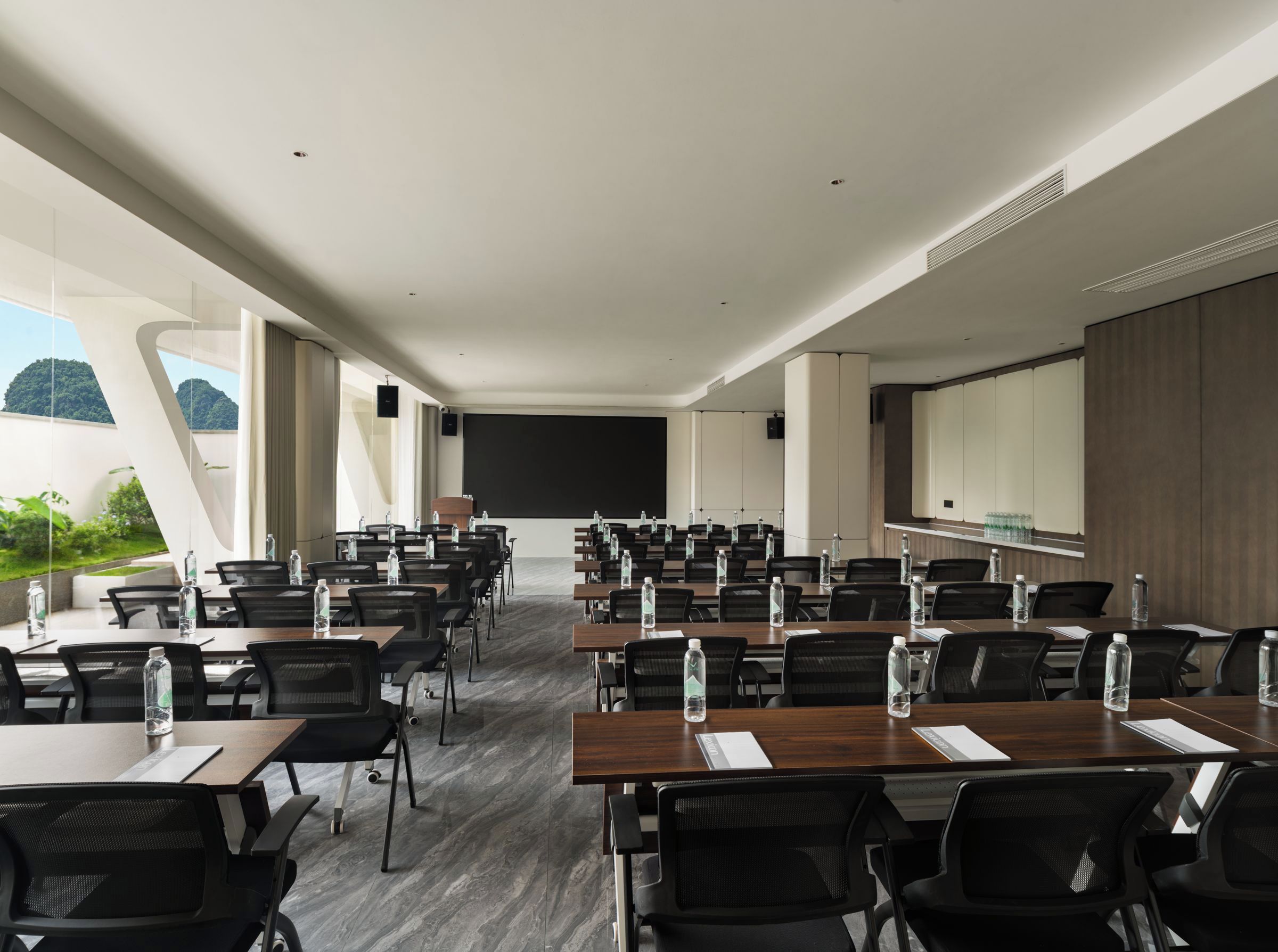 Sala de conferências com várias mesas e cadeiras, uma tela de projetor e uma janela grande com vista para o verde.
