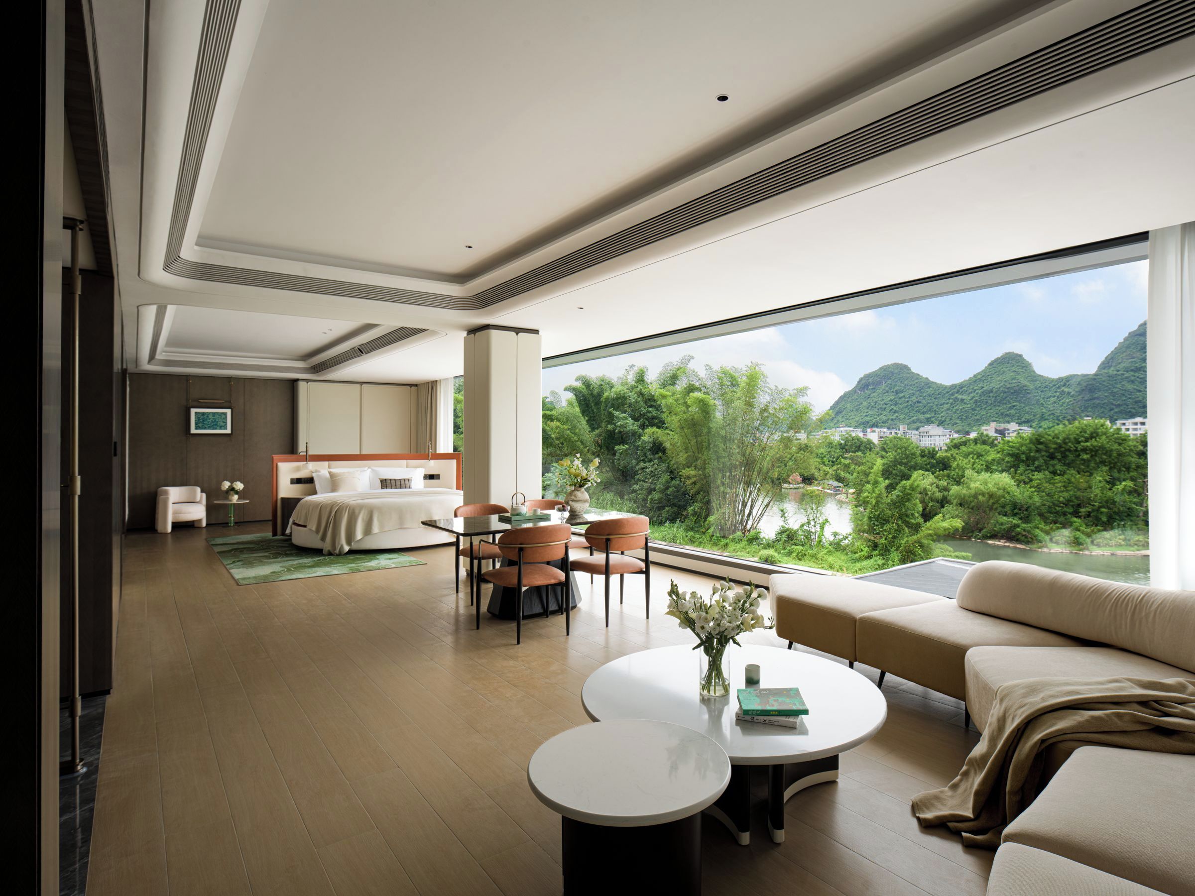 A modern hotel room with a large window, a bed, a seating area with a table and chairs, and a view of a river and mountains.