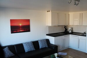A modern living space with a black sofa, a painting of a sunset, and a kitchen with white cabinets and a black countertop. - Holiday apartment with 1 bedroom (Westerland)