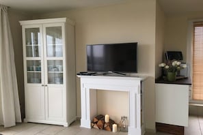 A living room with a television on a white console table, a cabinet with glass doors, and a window with blinds. - Holiday apartment with 2 bedrooms (Wangerland Horumersiel)