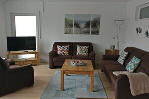 A living room with a sofa set, a coffee table, a television on a stand, and a painting on the wall. - 1 in the periwinkle house (Cuxhaven-Duhnen)