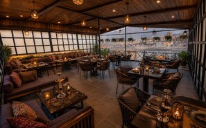 An indoor dining area with multiple tables and chairs, a large window offering a view of a cityscape, and hanging plants. - The Cove Hotels And Resorts  (Srinagar)