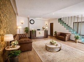 A modern hotel lobby with leather sofas, a reception desk, a clock, and a staircase with glass steps.
