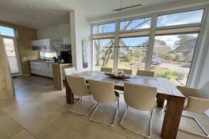 Una cocina moderna con armarios blancos, un comedor con mesa y sillas de madera, y ventanas amplias con vistas al exterior.