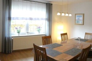 Une salle à manger avec une table en bois, quatre chaises, une fenêtre avec des rideaux, un radiateur et un luminaire fixé au mur.
