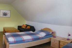 Un lit simple avec une couverture à carreaux, une petite table de chevet en bois avec une lampe, un canapé noir avec des coussins et une table en bois.