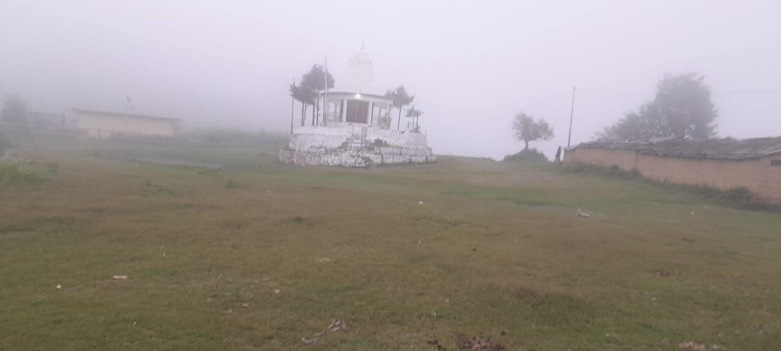 A foggy landscape with a small white building, possibly a gazebo, surrounded by grass and trees.