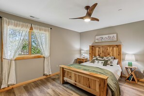 A bedroom with a wooden bed, a nightstand with a lamp, a ceiling fan, and a window with sheer curtains. - 5BR chalet with hot tub, fireplace, and wooded view (McHenry)