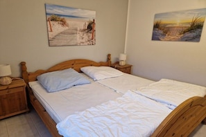 En dubbelsäng med vita sängkläder, en sänggavel i trä, två sängbord med lampor och två tavlor med motiv av strandscener, upphängda på väggen.