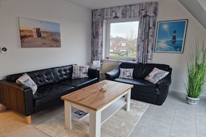 A living room with two black leather sofas, a wooden coffee table, and a large window with curtains. - Holiday apartment Strandglück (Hooksiel)