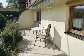 A patio with a round table and two chairs, a window with a lace curtain, and a small door. - at the country house Alte Schmiede (Rettenberg)