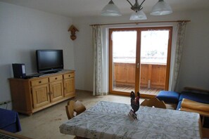 A room with a television on a wooden cabinet, a table with a patterned tablecloth, and a balcony with wooden flooring and blue cushions. - Holiday farmhouse with 2 bedrooms (Nußdorf)